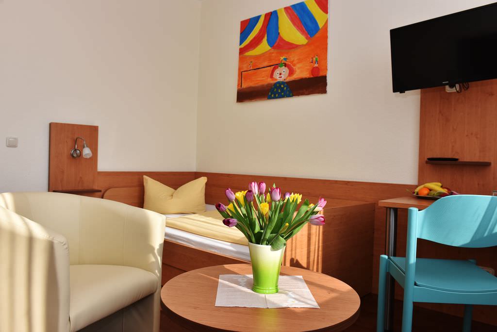 Vor Kopf ein Bett mit Kindergemälde eine Zirkusszene darüber, rechts davon ein kleiner Schreibtisch mit Fernseher darüber, links ein Sessel. in der Mitte im Vordergrund ein runder Tisch mit Blumenvase und Blumen.