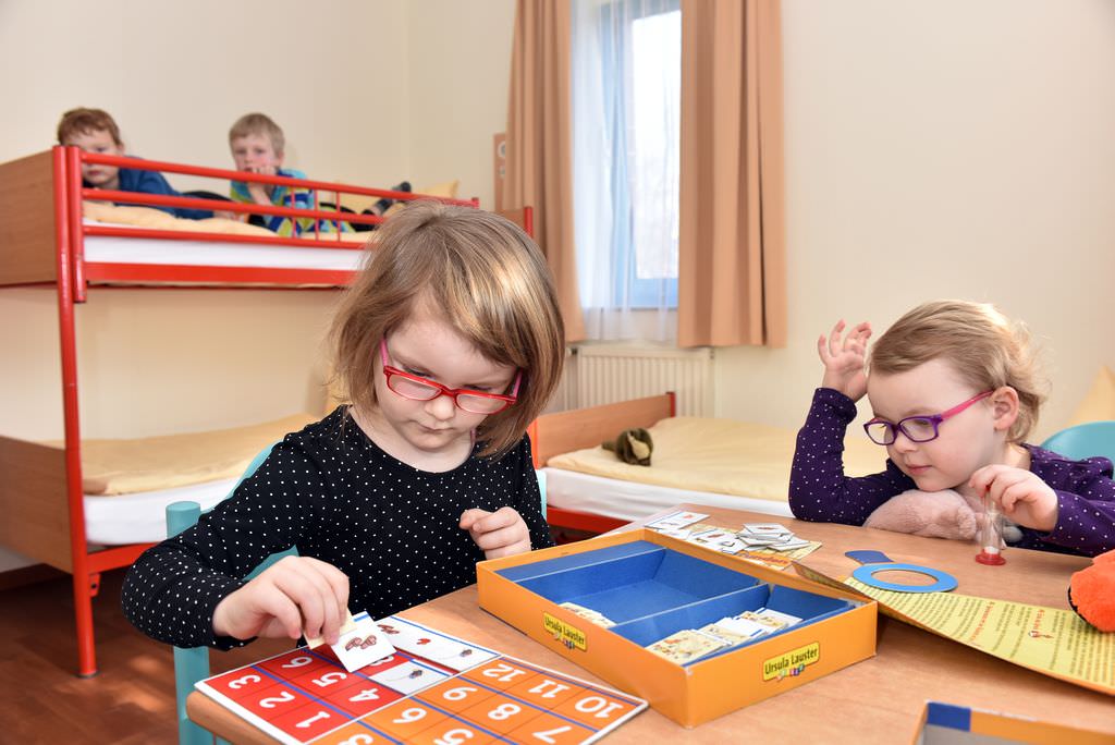 Zwei Mädchen spielen Memory an einem Tisch und werden dabei von zwei Jungen beobachtet, die in der oberen Etage eines Etagenbettes liegen.