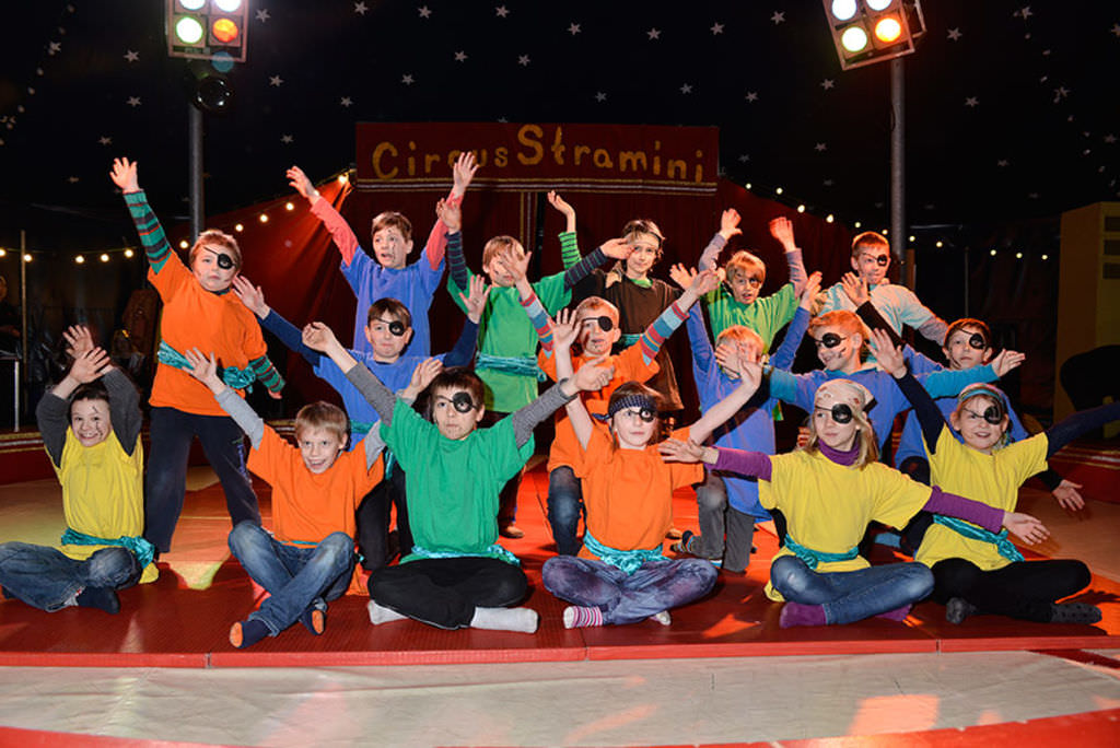 Eine Gruppe von Kindern in Piratenkostümen posieren mit den ausgestreckten Armen in einem Zirkuszelt. Im Hintergrund ist zu lesen: Circus Stramini.