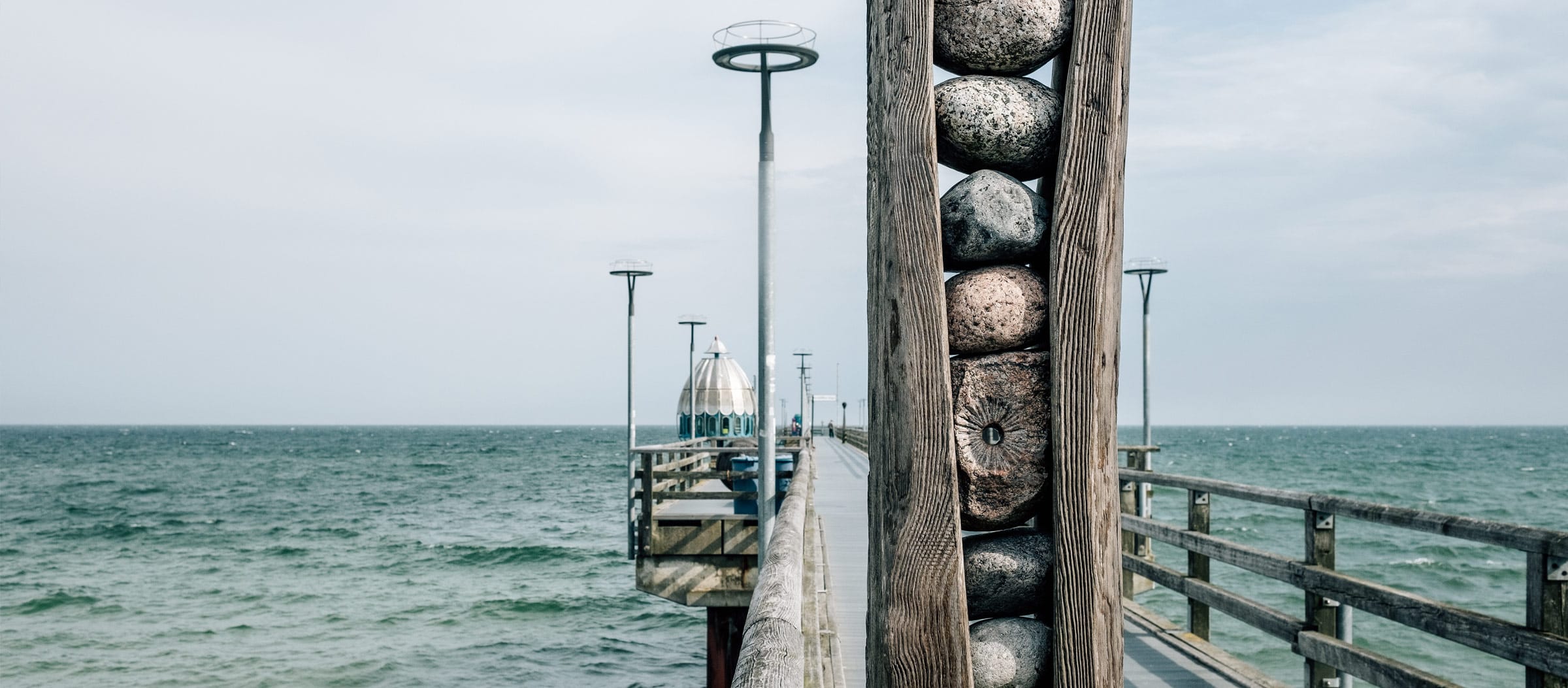Ein Holzsteg der hinaus auf die Ostsee führt, im Vordergrund eine gespaltener Holzpfahl mit Steinen in der Mitte, in einem Stein befindet sich ein rundes Guckloch.
