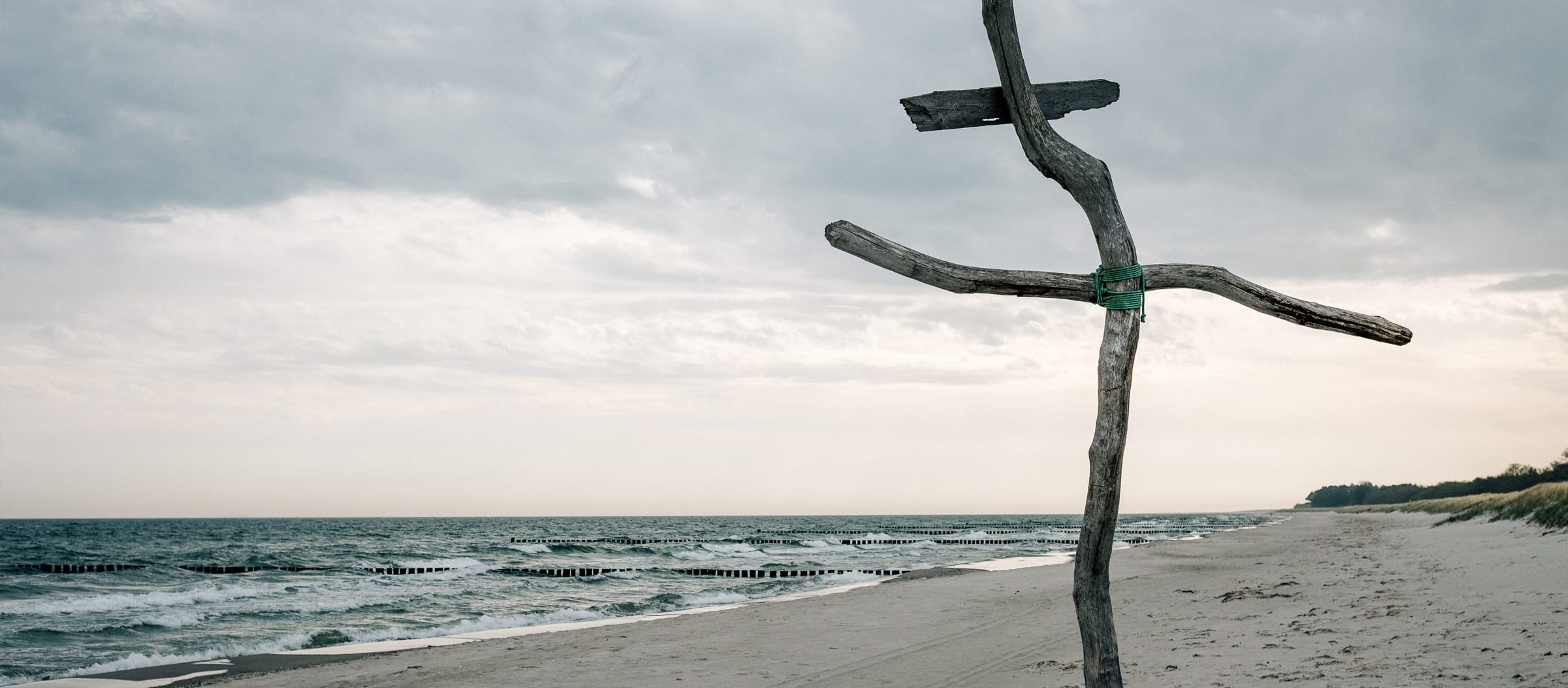 Unter bedecktem Himmel: links die etwas stürmische Ostsee, rechts der Sandstrand, im Vordergrund ein krummes Holzkreuz.