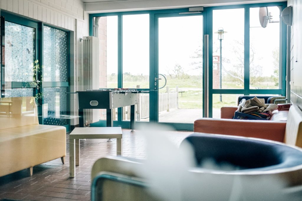 Freundlich gestalteter Aufenthaltsraum mit Sitzecken, Kicker und Blick durch bodentiefe Fenster und Terassentüre nach draußen.