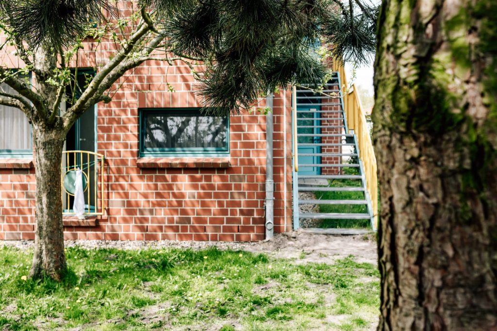 Rechts eine Treppe, die entlang der Fassade eines Backsteingebäudes zu einer Türe führt. Links eine Backsteinfassade mit einem Fenster und einem kleinen Balkon, davor ein Baum.