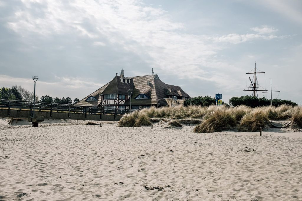 Ein weitreichender Sandstrand im Vordergrund, der in der Mitte der Düne in trockenes Gras übergeht. Dahinter ein großes Haus mit markantem Ziegeldach und Gauben. Der Himmel ist bewölkt und hell.